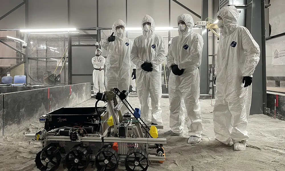 四名穿着实验室清洁服的学生聚集在地面上的一个多轮机器人周围.'